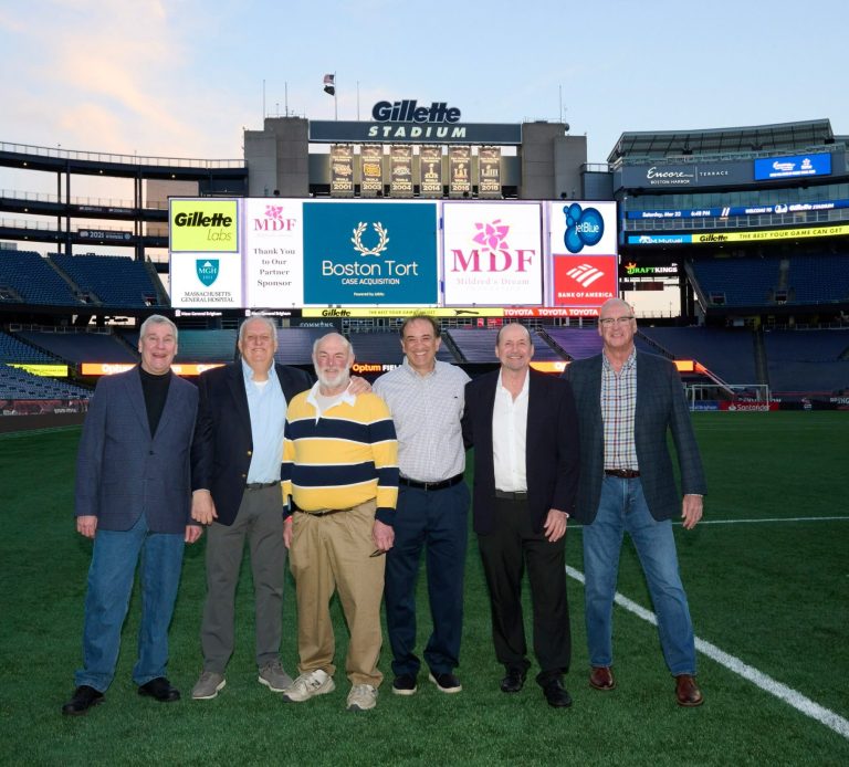 Group of six men standing on Gillette Stadium field in front of a large stadium screen for a charity event.
