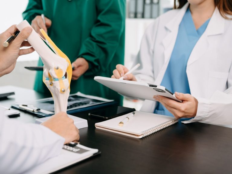 A medical consultation with professionals discussing a knee model and notes.