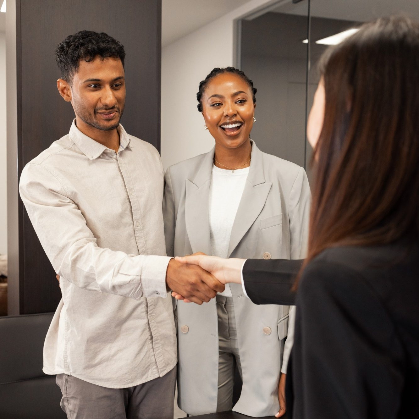 Two people shake hands while a woman smiles in a modern office setting.