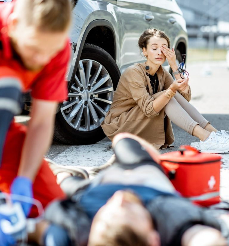 A woman sitting on the ground, visibly distressed, while paramedics attend to an injured person.